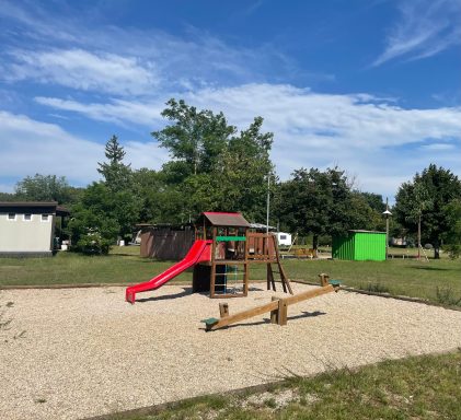 A aire de jeux avec toboggan rouge et balançoire sur un sol en gravier.