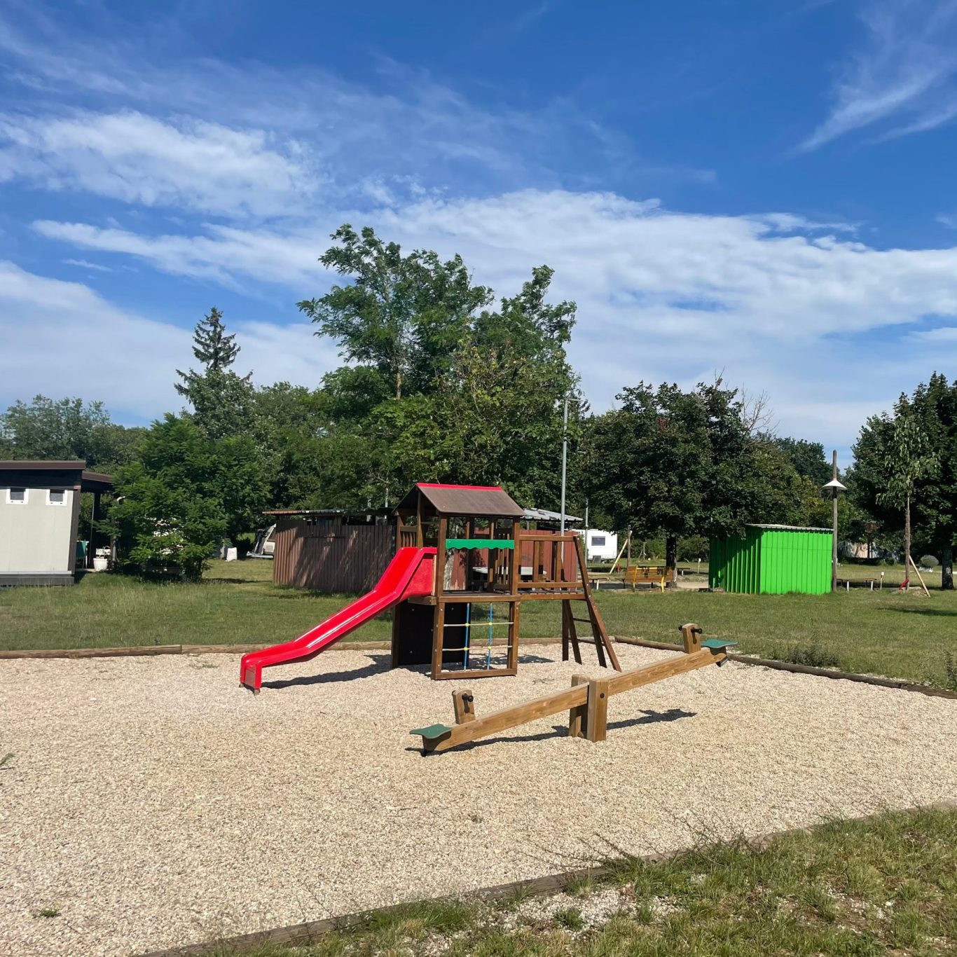 Aire de jeux avec toboggan rouge, balançoires et verdure environnante.